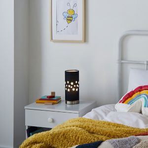 Navy table lamp with a cylindrical shape, featuring cut-out star patterns, sits on a white bedside table next to colorful books and a cozy yellow blanket.