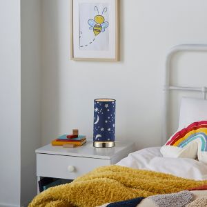 Silver table lamp featuring a navy blue cylindrical shade adorned with white moon and star patterns, placed on a bedside table beside a colorful blanket and framed artwork.