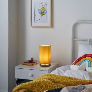 Silver hexagon table lamp with a warm glow, featuring a sleek design and fabric shade, placed on a bedside table next to colorful books and a cozy blanket.
