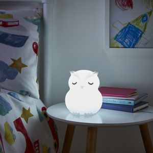 White owl-shaped table lamp with closed eyes, made of soft material, placed on a wooden side table beside colorful bedding and stacked books.