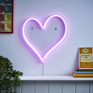 Pink neon heart wall light with a sleek design, mounted on a wall, featuring a white cord and surrounded by colorful decor and books on a shelf below.