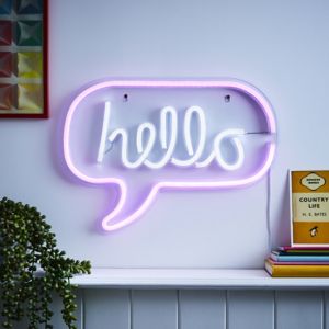 White neon wall light shaped like a speech bubble with the word "hello" in cursive, outlined in pink, mounted on a wall with colorful artwork and books nearby.