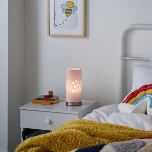A pink cylindrical table lamp with heart cutouts, resting on a bedside table beside a colorful striped pillow and a cozy yellow throw blanket.