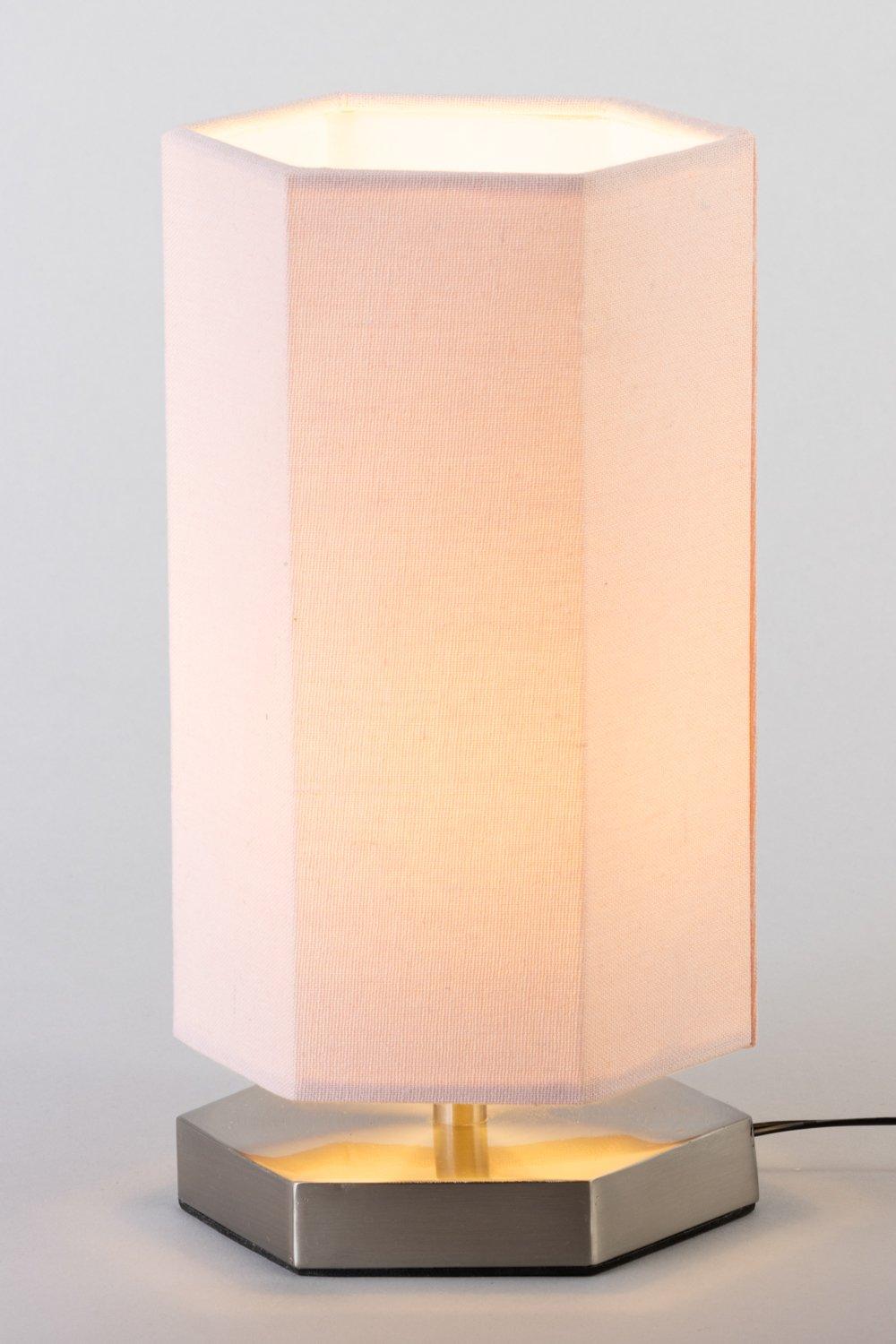 Silver hexagon table lamp with a soft pink fabric shade, featuring a warm glow from the interior light, set on a sleek metallic base.