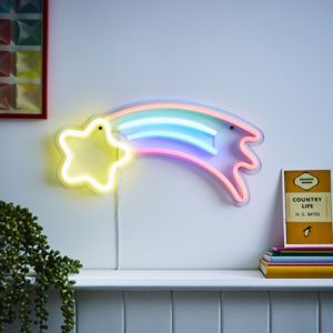Colorful neon wall light shaped like a shooting star with a yellow star and pastel rainbow stripes, mounted on a white wall above a shelf with books and a plant.