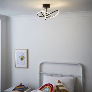 Black flush ceiling light with a whirly design, featuring sleek, curved LED strips, illuminating a cozy bedroom with a bed, colorful blanket, and framed artwork on the wall.