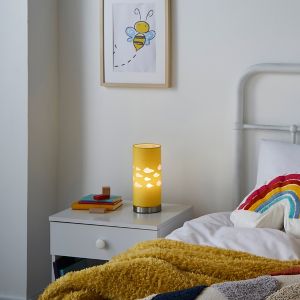 Yellow cylindrical table lamp with a cloud pattern, metallic base, placed on a bedside table next to colorful books and a cozy blanket in a bright bedroom setting.