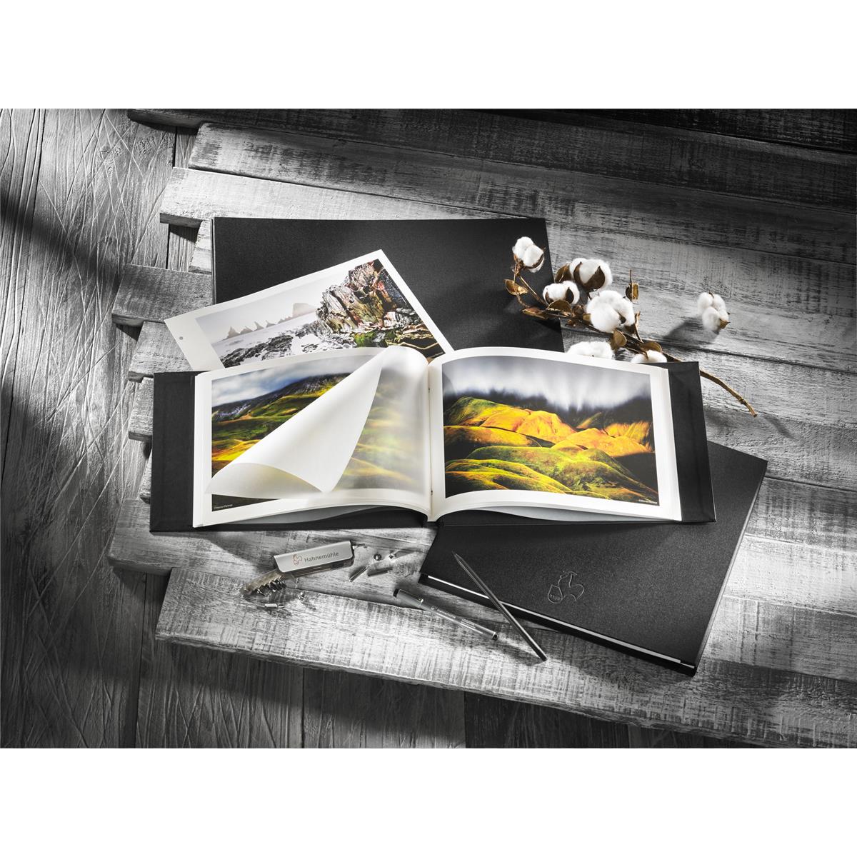 Black linen album cover set by Hahnemühle, featuring an open album with vibrant landscape photos, surrounded by cotton and tools on a textured wooden surface.