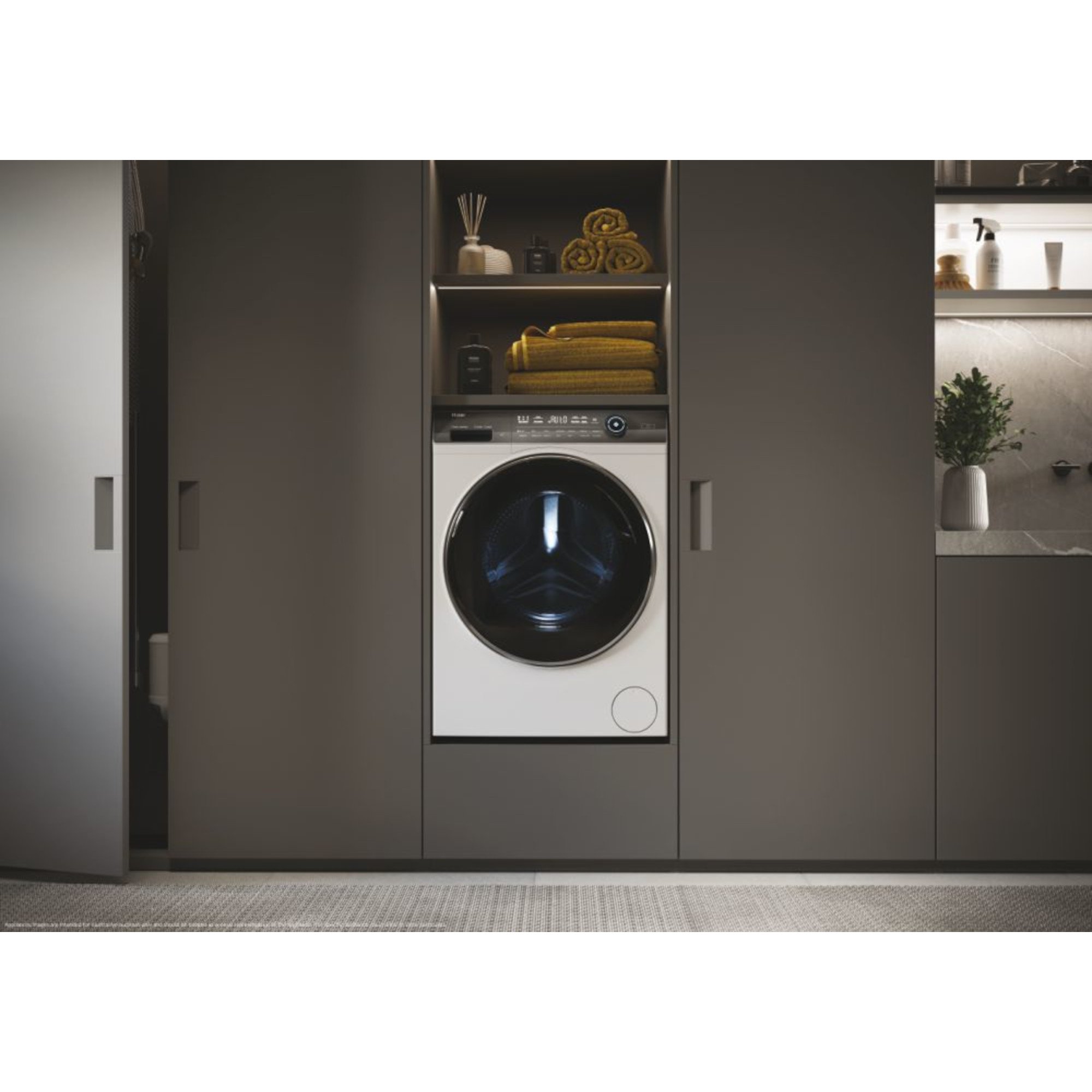 Front-loading washing machine in sleek white finish, featuring a large black door, digital control panel, and positioned within modern cabinetry alongside neatly stacked yellow towels.