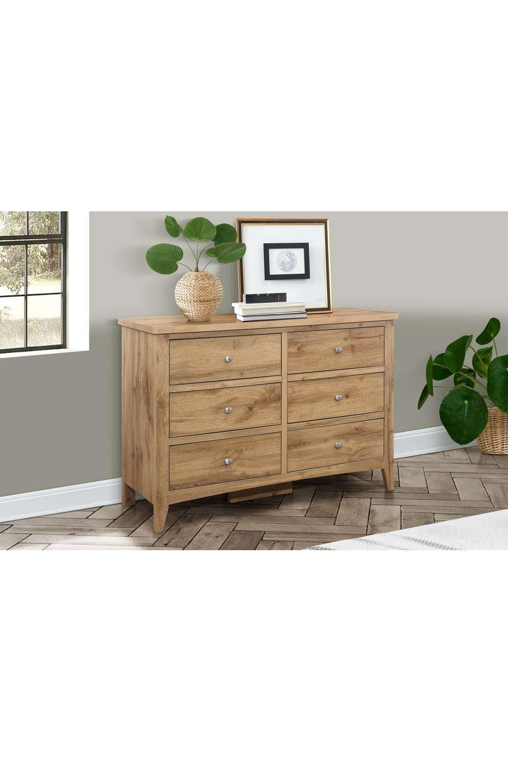 Birlea Hampstead 6 Drawer Chest features a warm brown oak finish, six spacious drawers with silver knobs, and a sleek design, complemented by decorative plants and artwork.