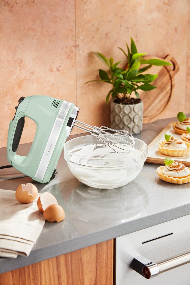 Pistachio green KitchenAid 5KHM9212BPT hand mixer with stainless steel beaters beside a glass bowl of cream, eggs, and pastries on a kitchen countertop.