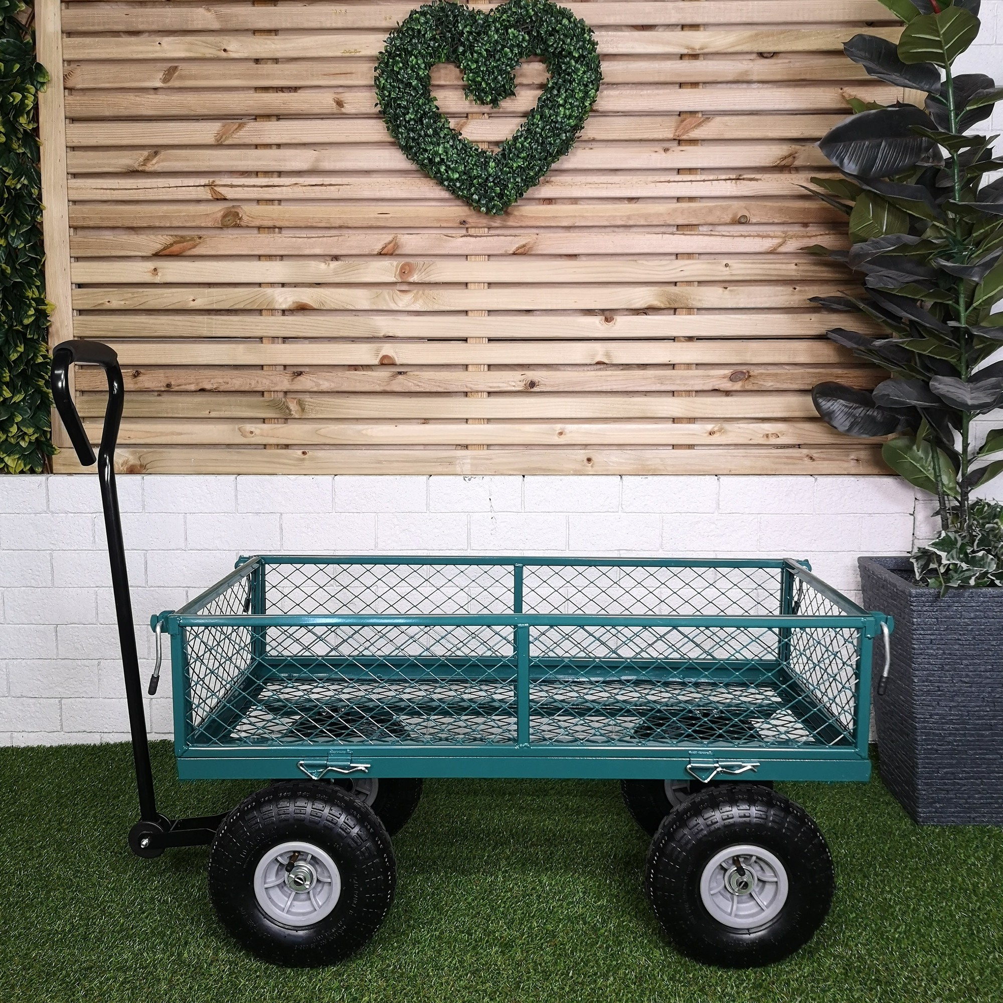 Heavy-duty green metal garden cart with a mesh design, sturdy wheels, and a black handle, set against a wooden backdrop with greenery.
