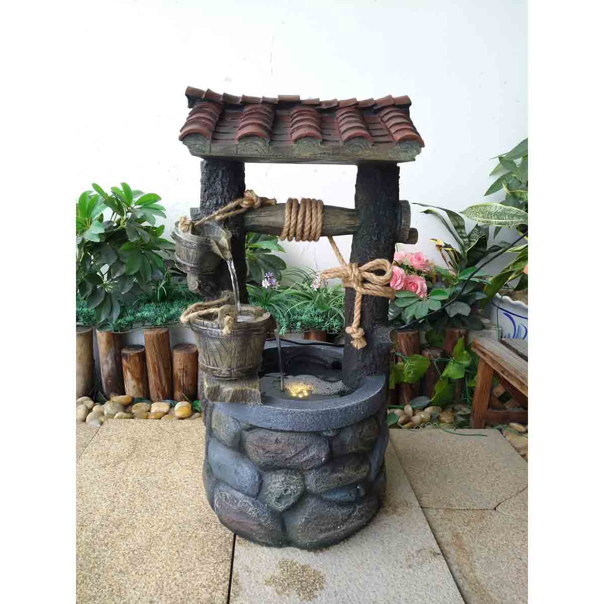 Grey stone-like wishing well water feature with a rustic wooden roof, rope accents, and an LED light, surrounded by greenery and decorative stones.