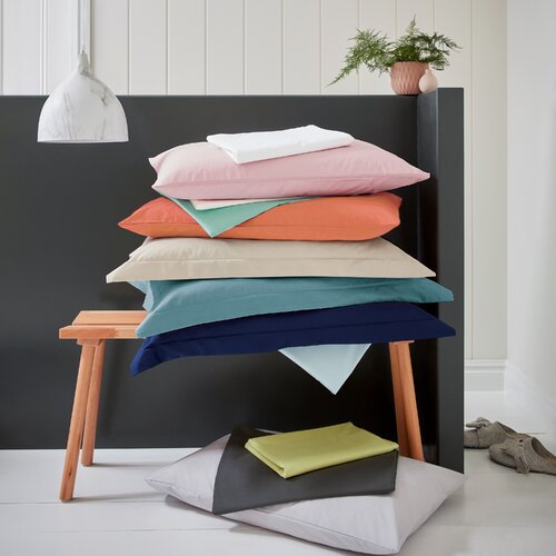 Light pink polycotton flat sheet from Helena Springfield, displayed among various colored pillows on a wooden stool against a dark wall, showcasing its soft texture and super king size.