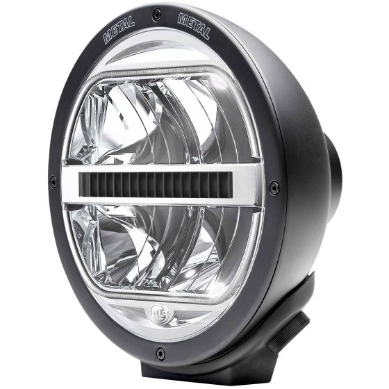 Round Hella Luminator LED lamp in black metal, featuring a clear lens with dual reflective surfaces and a side-mounted adjustment knob for directional light control.