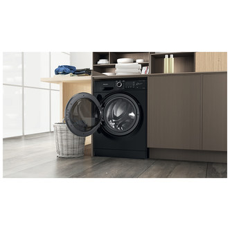 Black Hotpoint NDB9635BSUK washer dryer with a transparent door, showcasing a sleek design, placed in a modern kitchen setting with wooden cabinets and a laundry basket.