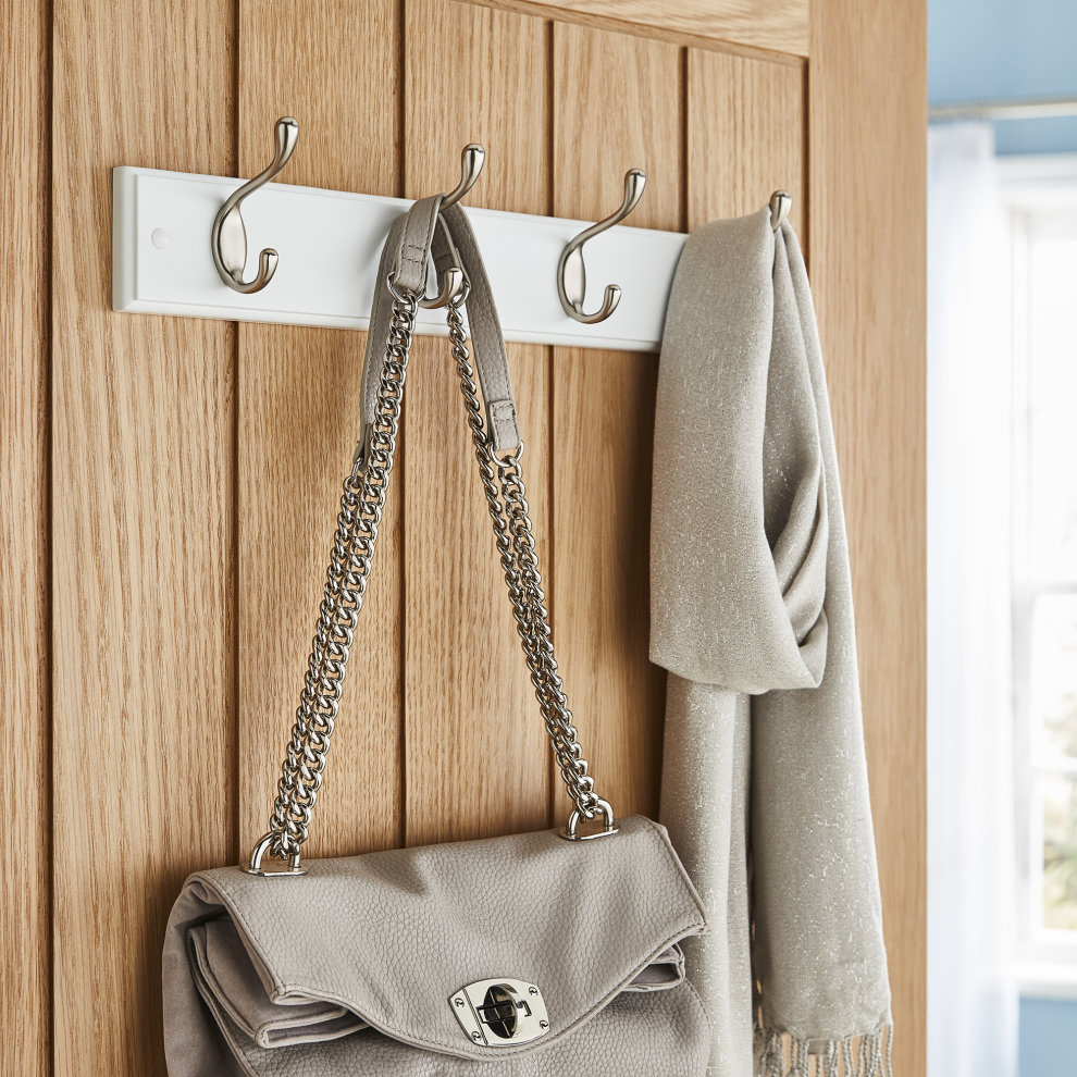 Four double coat hooks in polished chrome mounted on a white wooden board, holding a gray handbag and a light gray scarf, set against a wooden door backdrop.