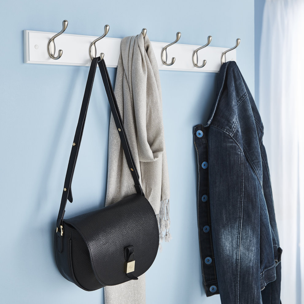 Six double coat hooks in a polished silver finish mounted on a white wooden board, with a black handbag, gray scarf, and denim jacket hanging on them against a blue wall.