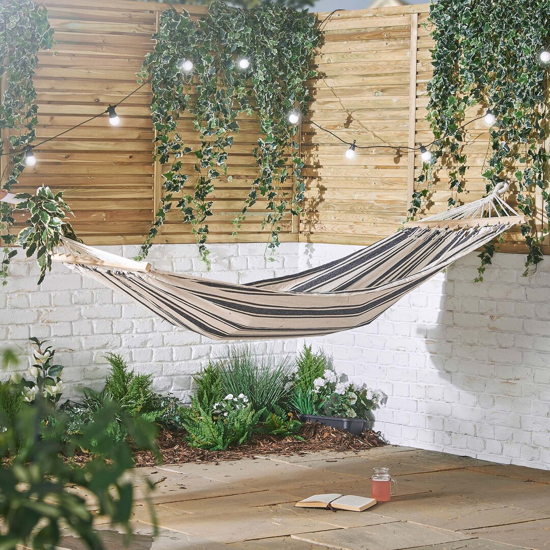 Striped cotton hammock in beige and navy, suspended between wooden posts, surrounded by greenery and fairy lights, with a book and drink on a stone floor below.