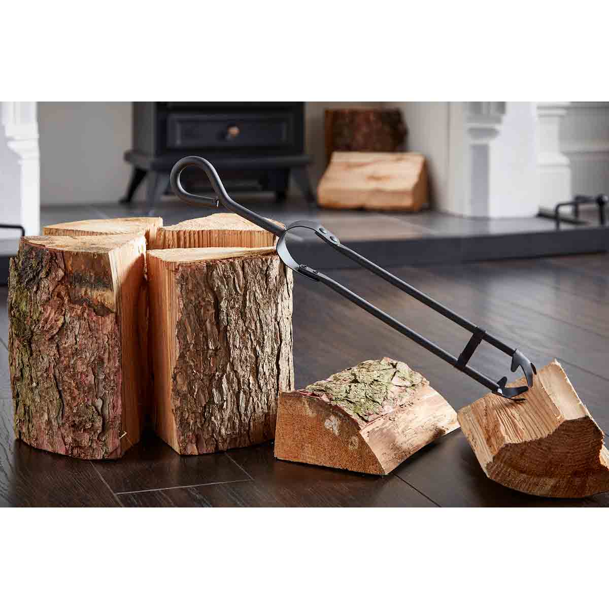Black metal tweezer fire tongs with a curved handle, positioned next to stacked logs with rough bark, set against a wooden floor and a fireplace backdrop.