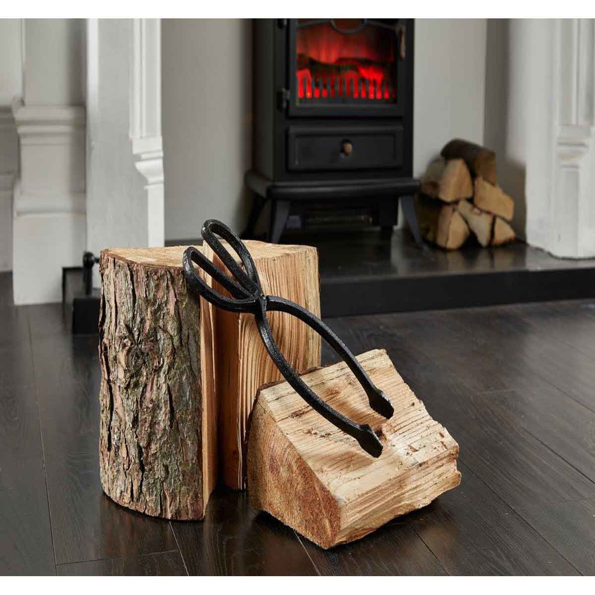 Matt black heavy metal tongs with a scissor design resting on two wooden logs, set against a cozy backdrop with a stove and stacked firewood.