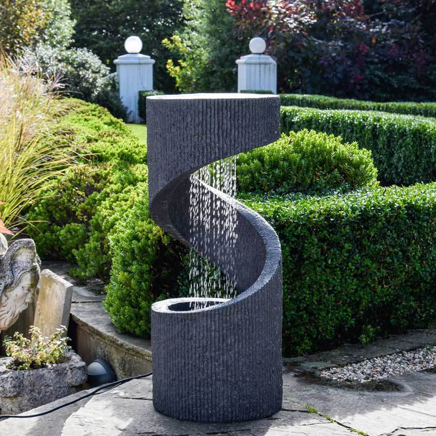 A sleek, dark granite water feature with a spiral design, featuring cascading water and surrounded by lush greenery and neatly trimmed hedges.
