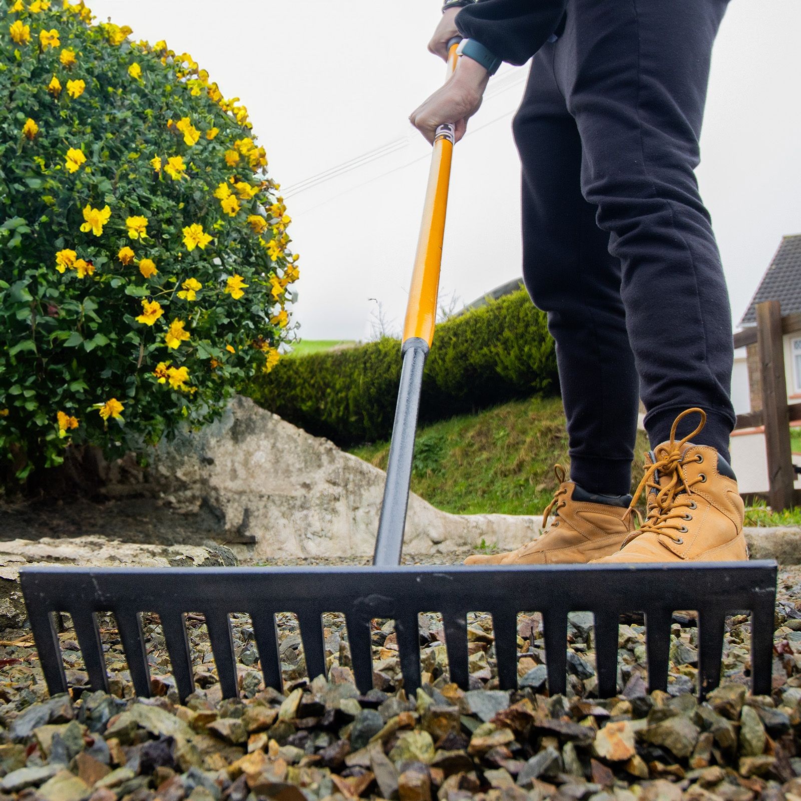 JCB Industrial Contractors Rake with 16 steel teeth, featuring a yellow handle and a black metal head, used for landscaping, held by a person wearing brown boots.