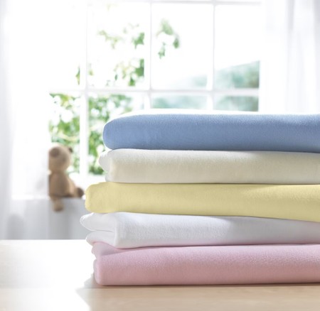 Soft jersey cot fitted sheets in blue, white, yellow, and pink stacked neatly, with a blurred teddy bear and window in the background.