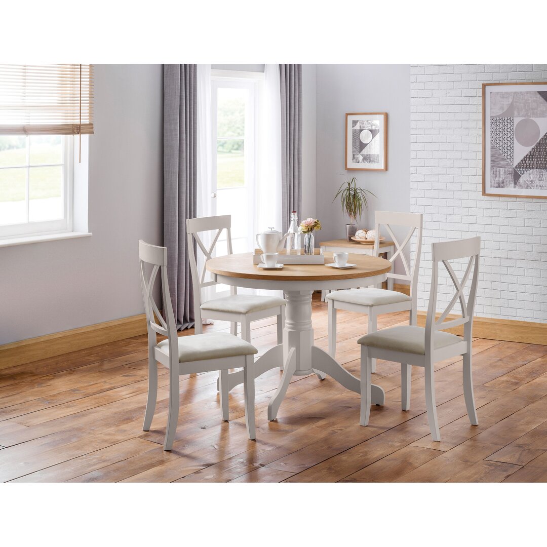 Round wooden dining table with a natural finish, surrounded by four white wooden chairs featuring an X-back design, set in a bright, airy room with light-colored walls and wooden flooring.