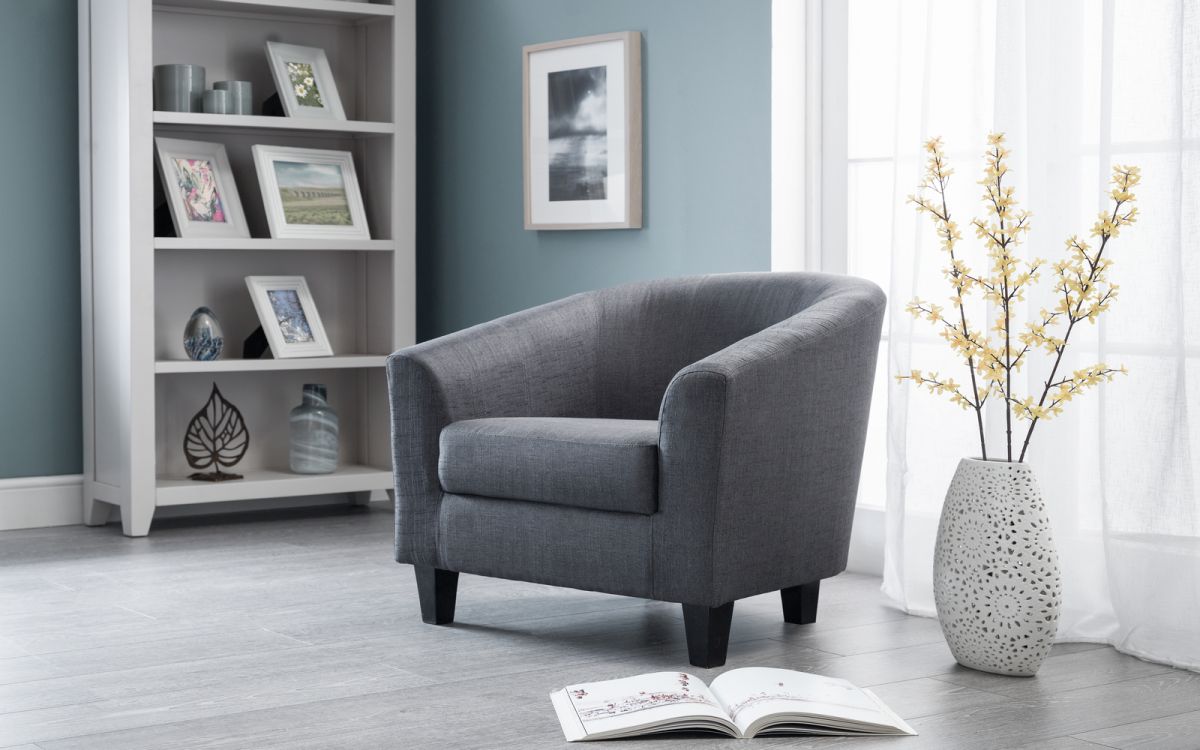 Slate grey tub chair with a curved back and upholstered fabric, featuring black wooden legs, placed in a bright room with decorative shelves and a vase of flowers.