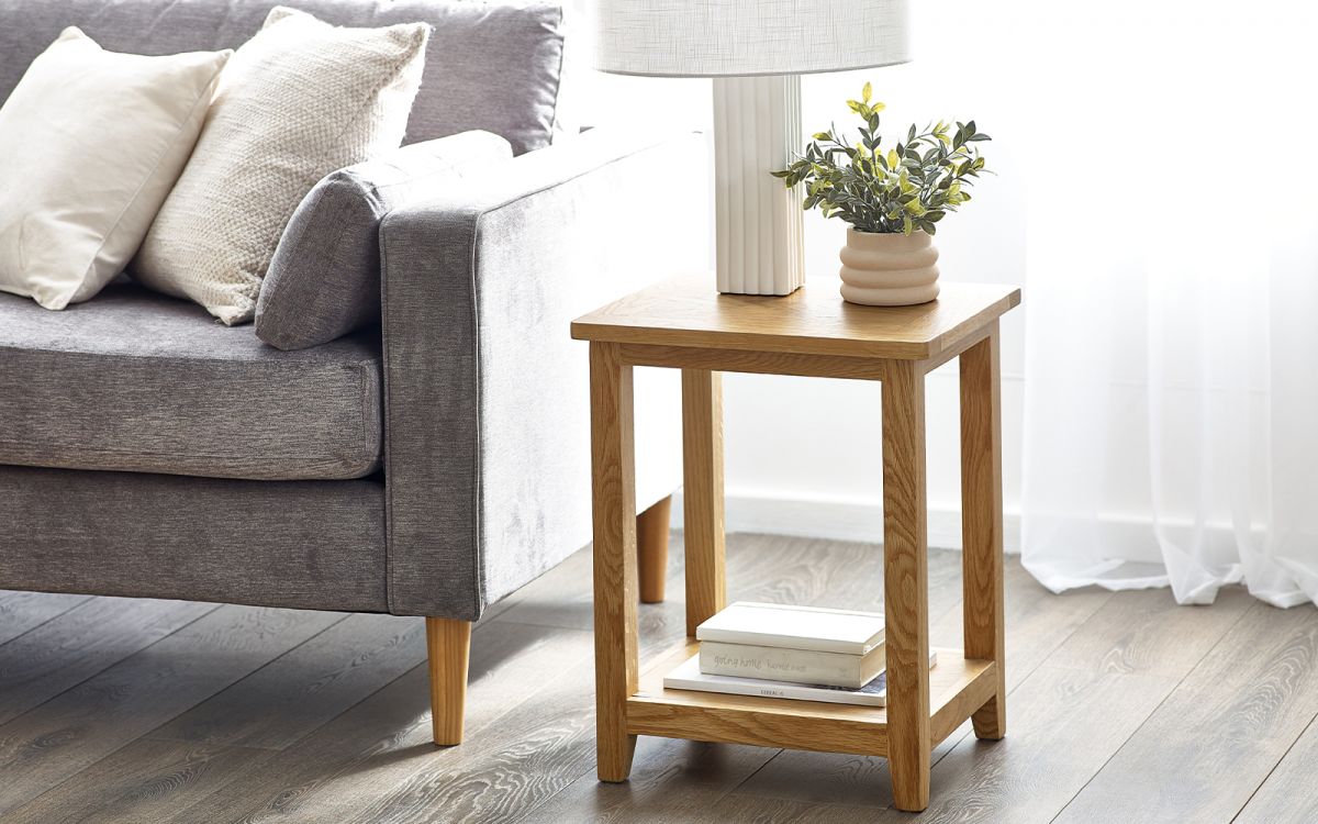 Light wood lamp table with a square top, featuring a lower shelf, positioned beside a gray sofa, adorned with a lamp and a potted plant.