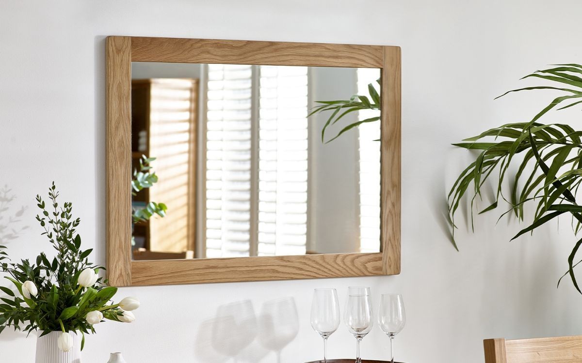 Rectangular wall mirror with a natural oak frame, featuring a simple design, reflecting light and greenery from nearby plants, enhancing a bright room atmosphere.