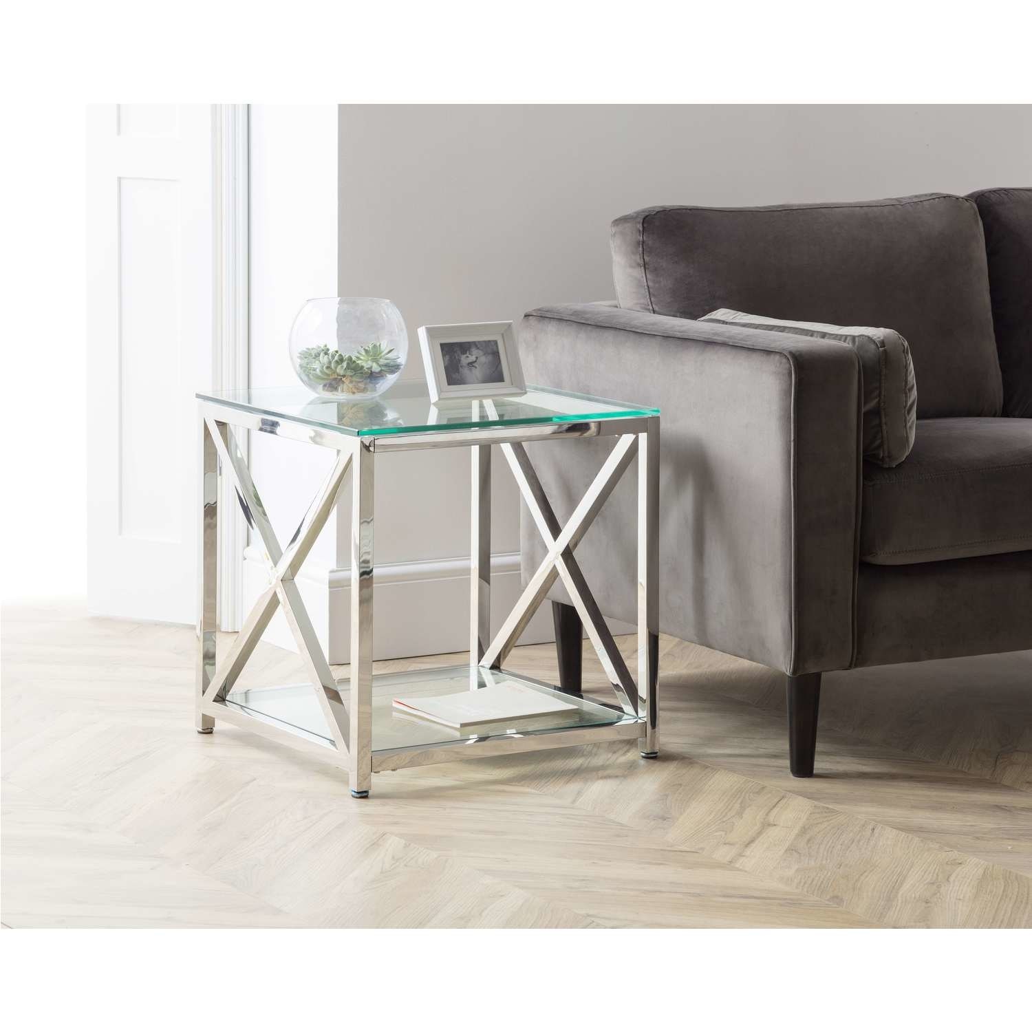 Stylish glass and silver lamp table with a 55x55x55cm frame, featuring an X-shaped design, placed beside a grey sofa, with decorative items on top.