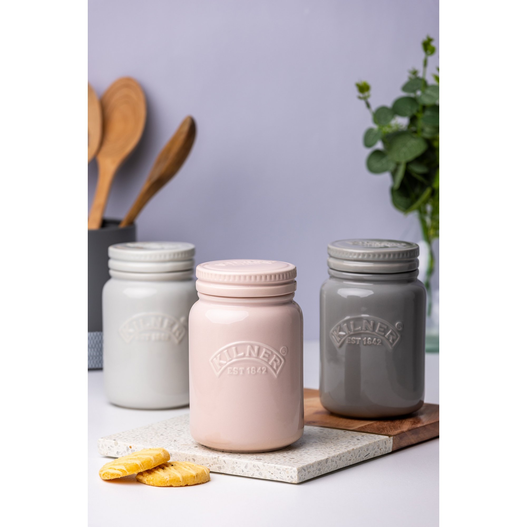 Kilner's 600ml ceramic storage jar in dusty pink features a push top lid, alongside grey and dark grey jars, set on a stone surface with kitchen utensils in the background.