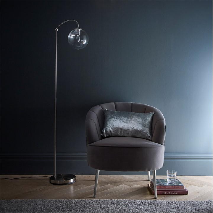 Satin nickel floor lamp with a ribbed glass shade stands beside a plush gray armchair adorned with a silver cushion, set against a dark wall.