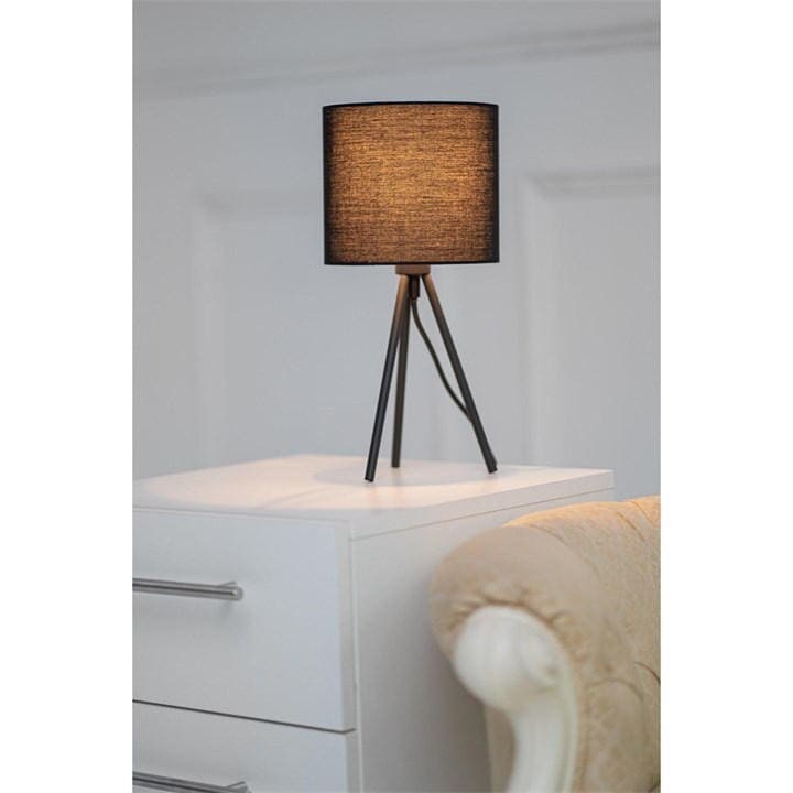 Small black tripod table lamp with a textured fabric shade, supported by three slender metal legs, placed on a white nightstand beside a light-colored upholstered chair.
