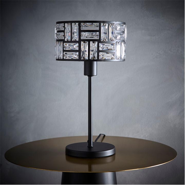 Black table lamp featuring an acrylic glass drum shade with a geometric design, mounted on a sleek black metal base, set against a textured gray background.