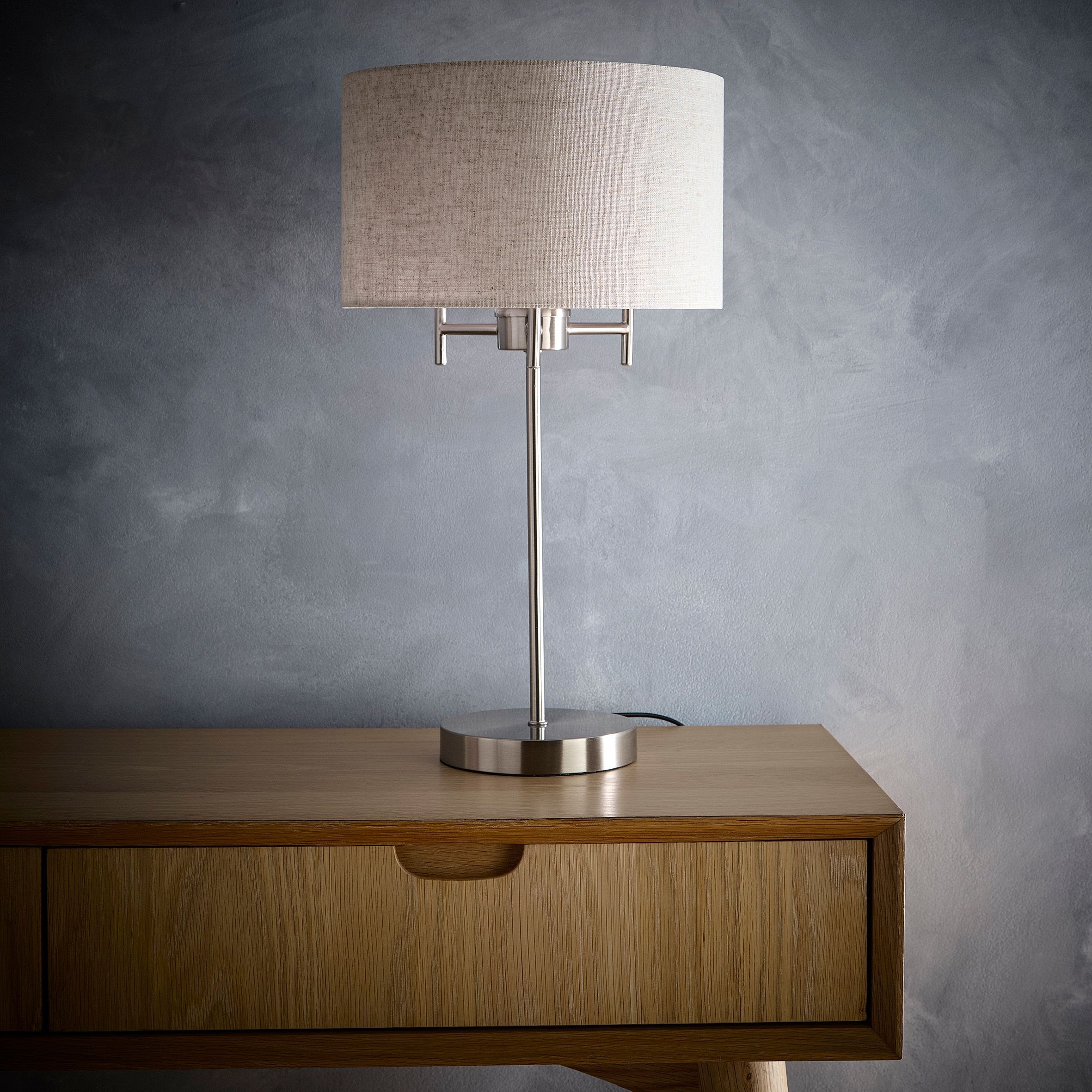 Tall table lamp featuring a satin nickel finish and a beige linen lampshade, set on a wooden table with a minimalist design.