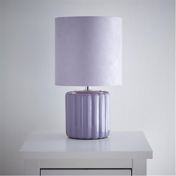 Lilac ceramic table lamp with a ribbed base and matching purple velvet shade, elegantly placed on a white bedside table against a light wall.
