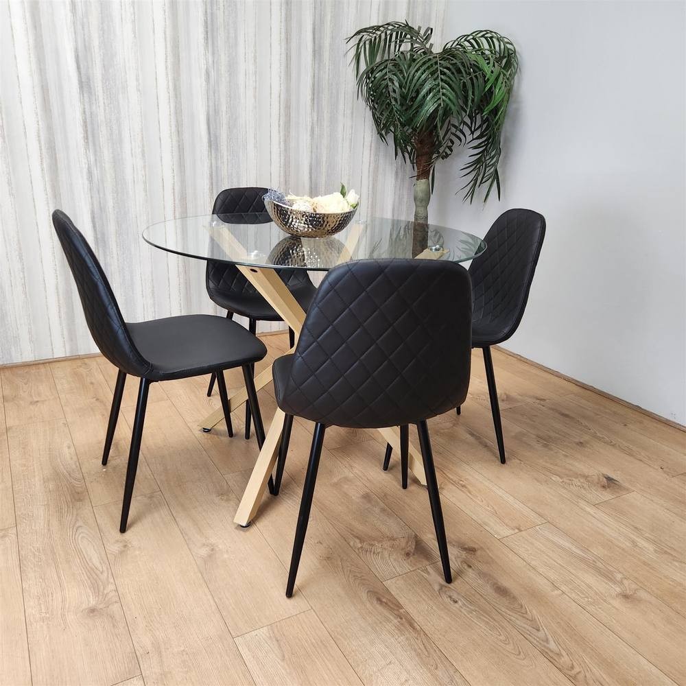Clear glass round dining table with a wooden base, surrounded by four black leather padded gem chairs featuring diamond stitching, set on a light wooden floor with a potted plant in the background.