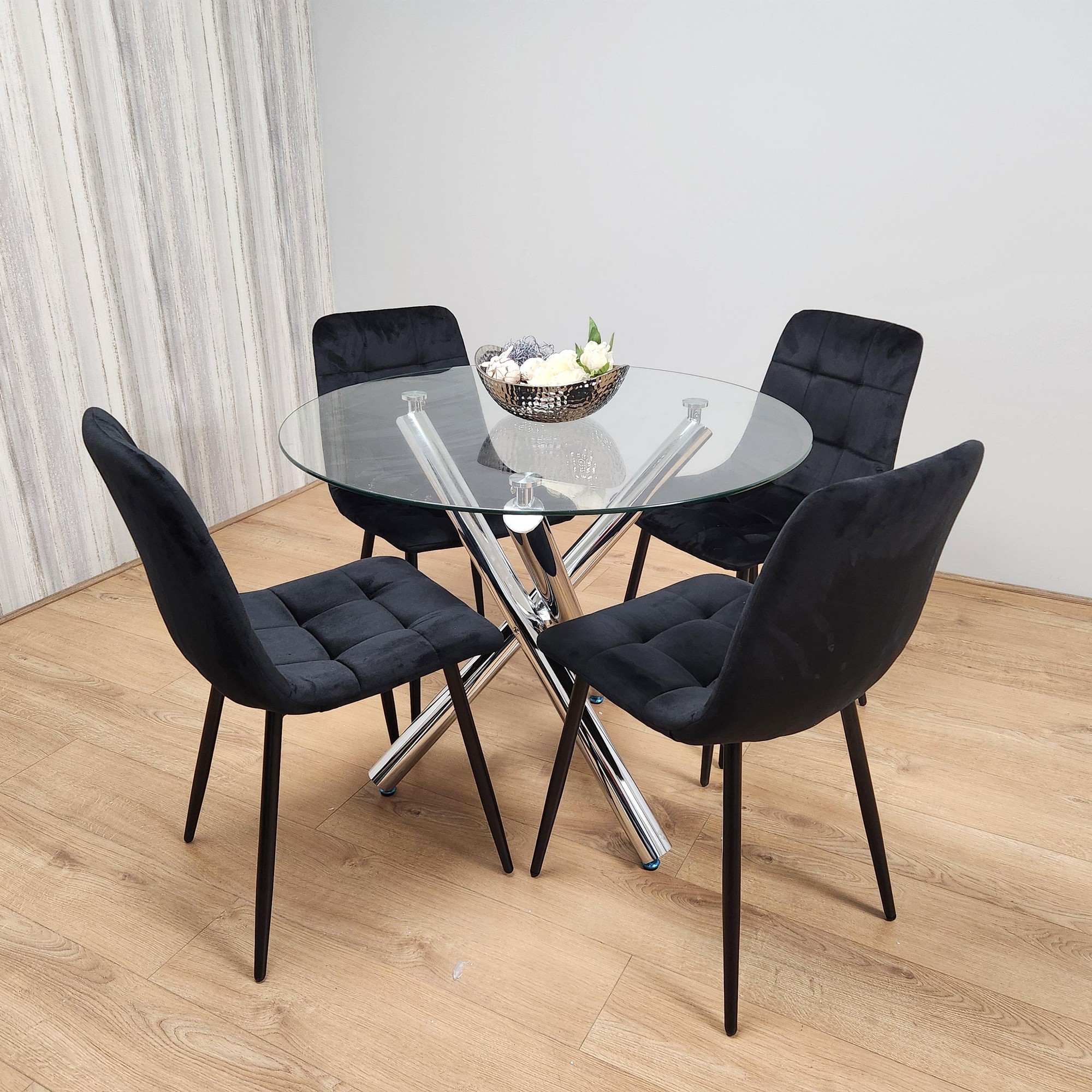 Round chrome metal dining table with a clear glass top, surrounded by four black tufted velvet chairs, set on a light wood floor.