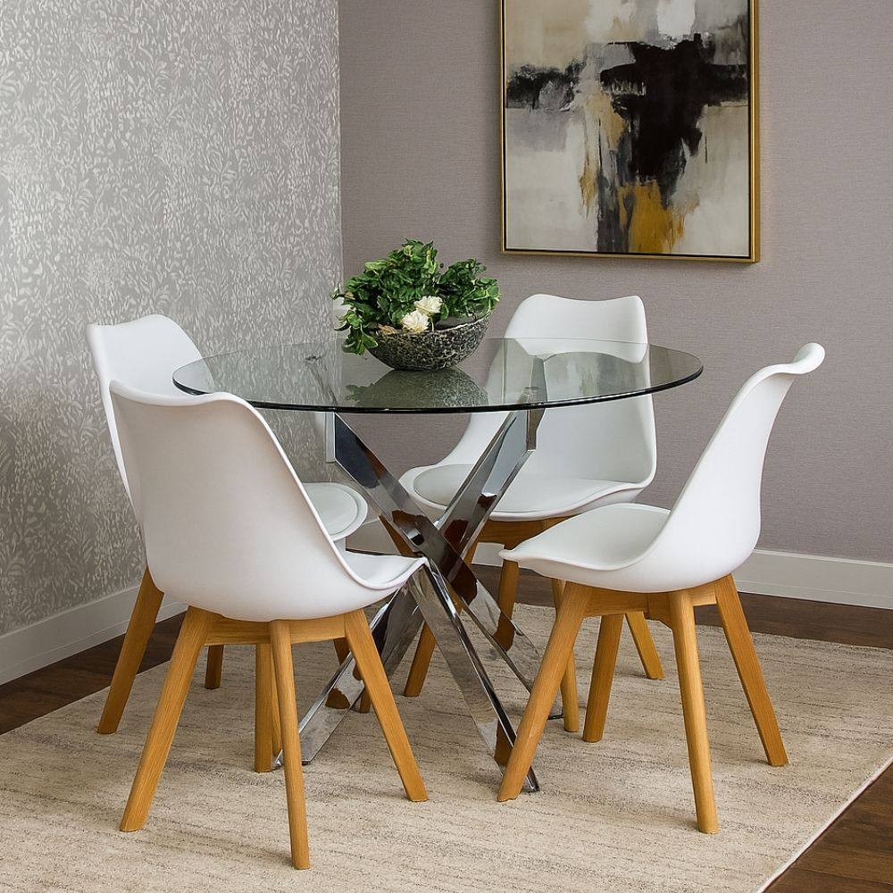 Round glass dining table with a chrome cross-base, surrounded by four white plastic chairs with wooden legs, set on a light-colored rug against a patterned wall.