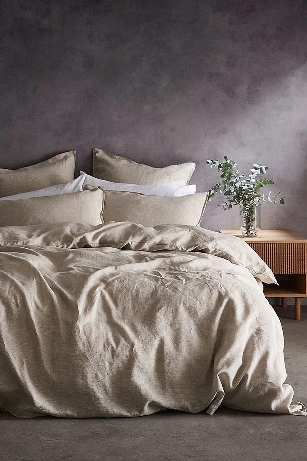 Soft beige linen duvet cover with a relaxed texture, accompanied by matching pillows and a wooden bedside table with a vase of greenery, set against a muted gray wall.