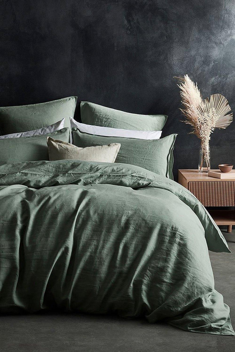 Sage green linen duvet cover set with matching pillows, featuring a textured finish, styled with decorative cushions and a wooden bedside table in a dark, moody setting.
