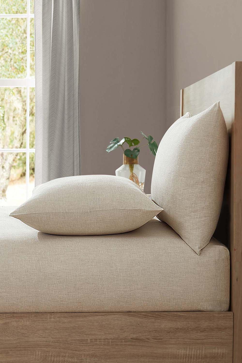 Light beige linen fitted sheet on a super king bed, paired with two matching pillows, set against a softly lit room with a window and greenery in the background.