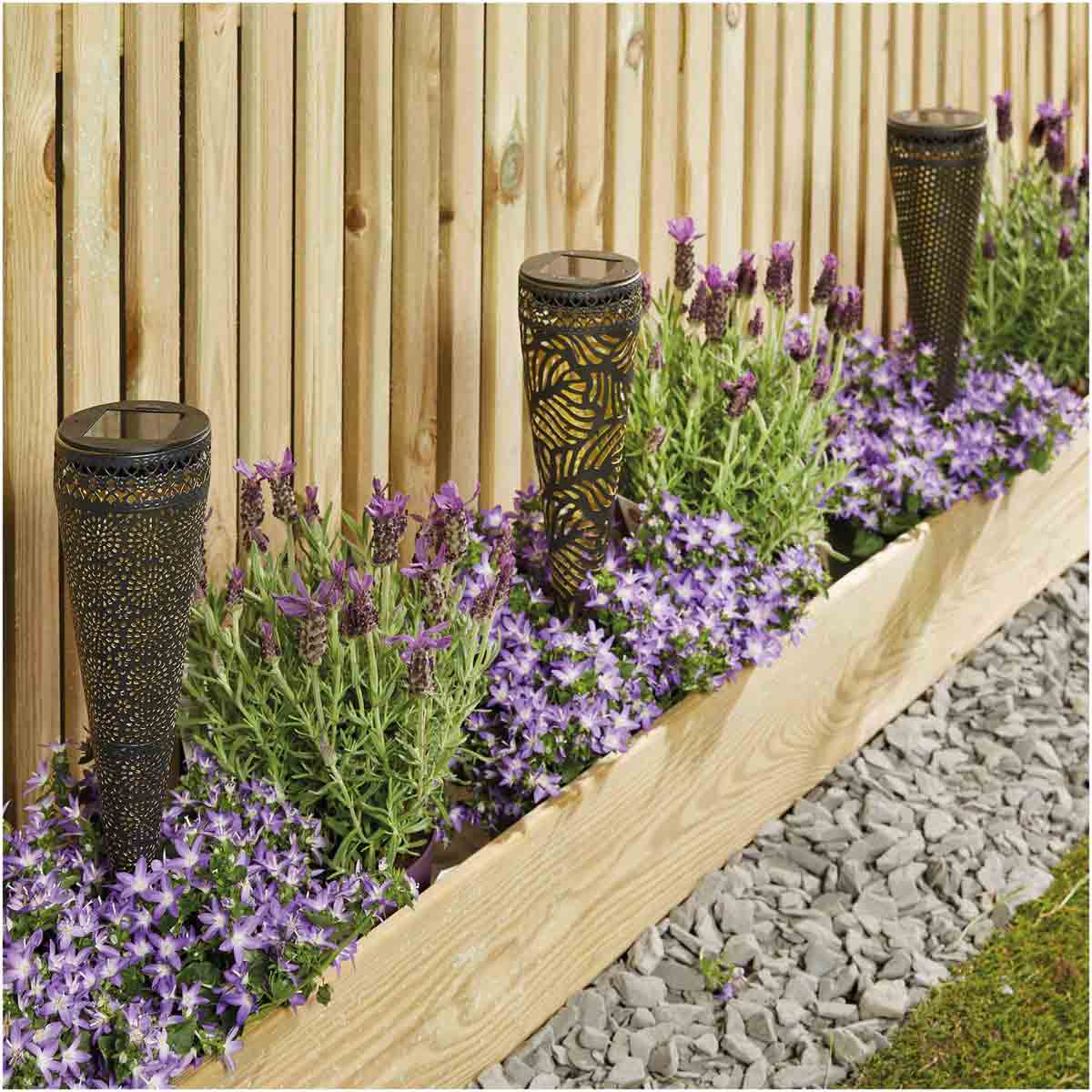Three decorative solar lights with a leaf pattern, set in a wooden planter filled with purple flowers and greenery, against a wooden fence backdrop.