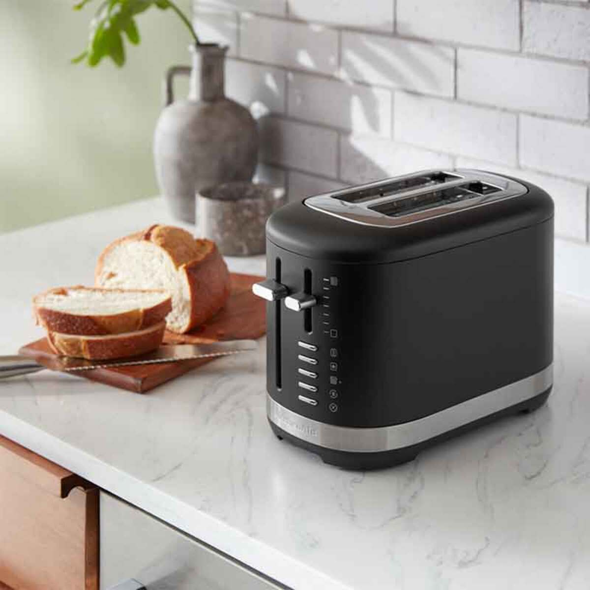 Matte black KitchenAid 5KMT2109BBM manual control 2-slice toaster with a stainless steel base, featuring a lever and multiple browning settings, next to sliced bread on a wooden cutting board.