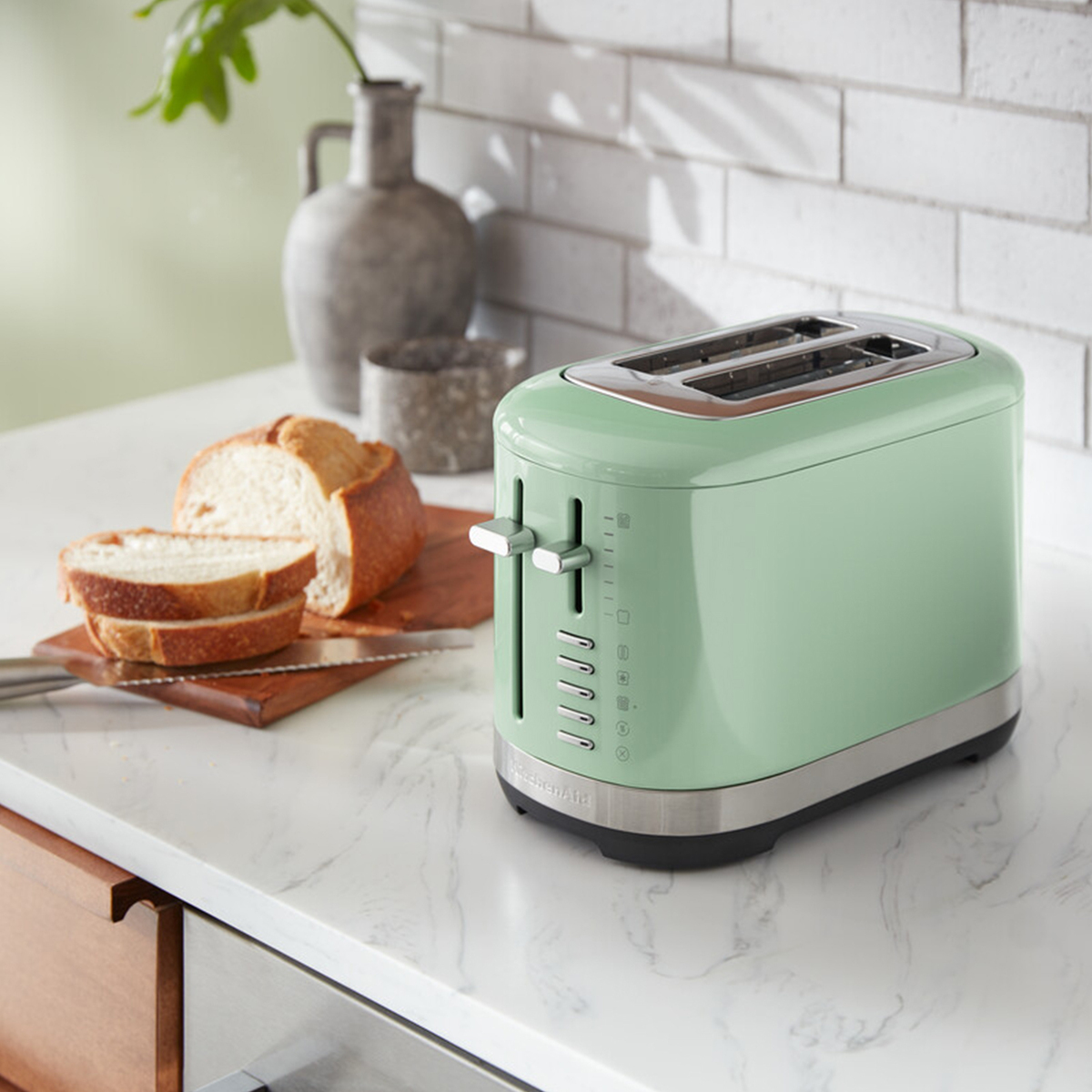 Pistachio green KitchenAid 5KMT2109BPT manual control 2-slice toaster with stainless steel accents, placed on a marble countertop beside sliced bread.