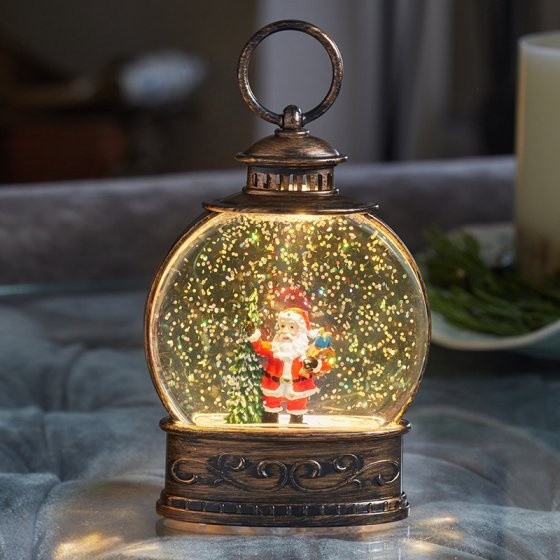 A warm white lantern featuring a festive Santa figurine inside a glass dome, surrounded by a snowy scene, with an ornate bronze base and a decorative handle on top.