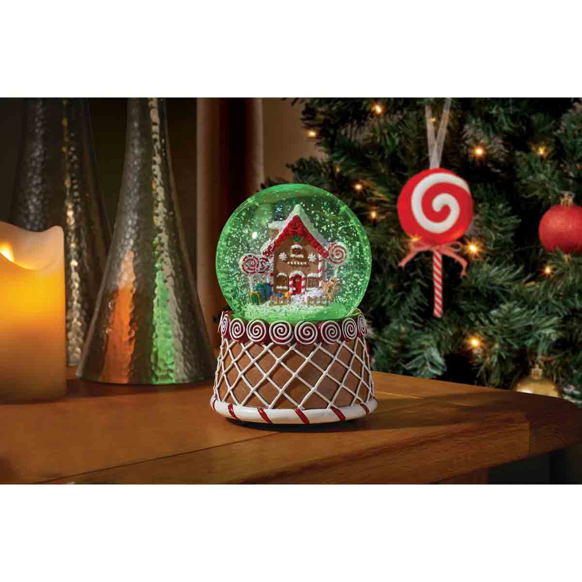 A 10cm musical gingerbread house snow globe features a colorful gingerbread house with candy details, set on a decorative base, surrounded by festive decorations.